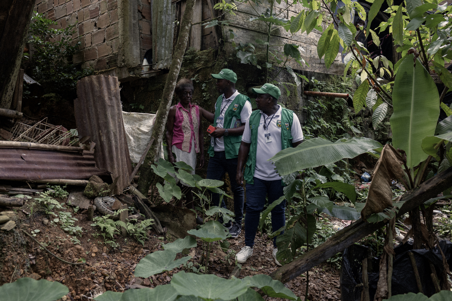 Deux bénévoles vêtus de gilets verts rendent visite à une femme âgée à l'air fragile dans sa cabane en bordure de jungle