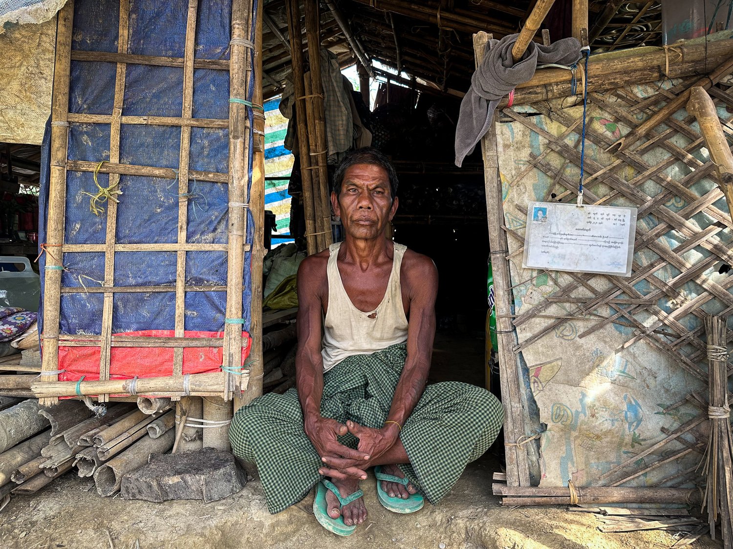 A man sits cross-legged in the doorway of a bamboo shelter.