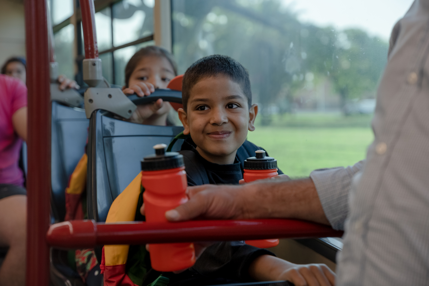 A boy smiles while sitting on a bus holding a water bottle.