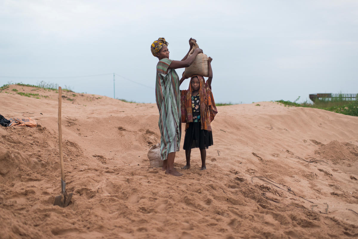 A displaced Congolese woman helps a girl balance a bag of sand on her head near Lake Tanganyika.