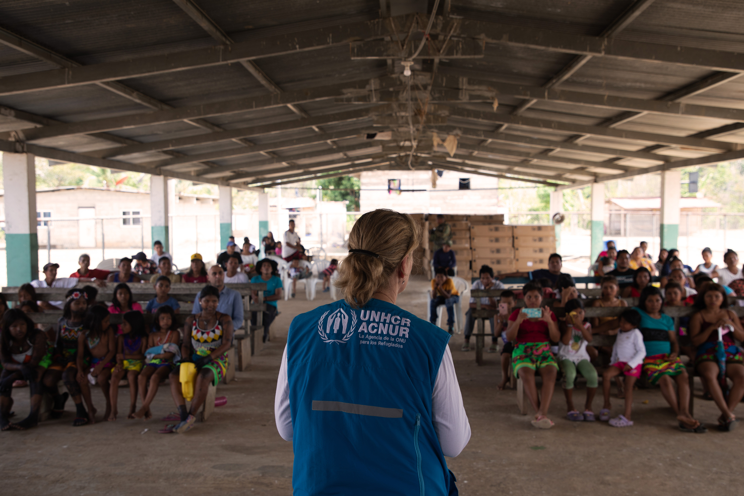 A woman in a branded UNHCR vest addresses a group of people in an open-sided building.