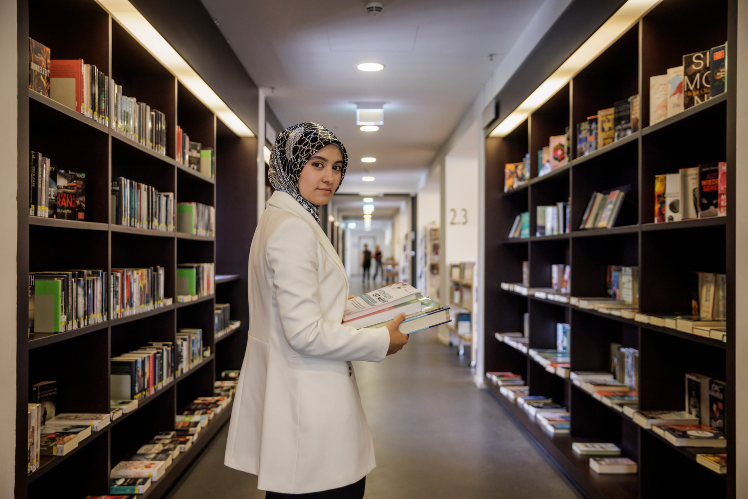 A woman in a lab coat stands between stacks of books in a library.
