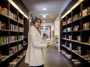 A woman in a lab coat stands between stacks of books in a library.