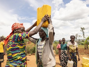 Women in colourful clothing, in a dry landscape, collect water in cannisters, carrying them on their heads or in their hands. 