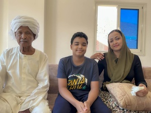 A boy sits on a sofa between his grandfather and mother in their apartment.