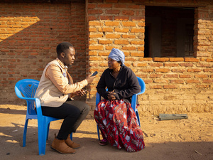 A man interviews a woman outside on two plastic chairs, in front of a brick building