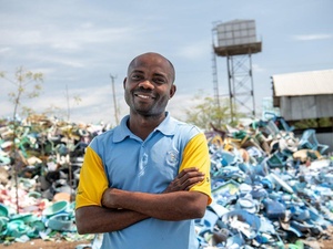 A smiling man stands in front of a huge pile of plastic waste.