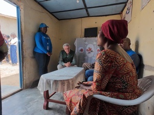 A woman seated on a plastic chair speaks to people seated and standing around a table inside a shelter