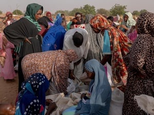 Outside in a sandy expanse with trees in the background, a large group of gather around sacks of grain on the floor