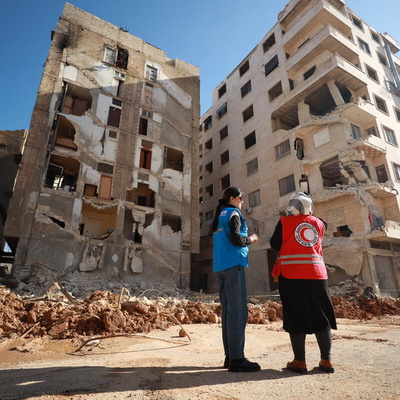 Une employée du HCR et une employée de la Croix-Rouge sont debout devant un bâtiment détruit.