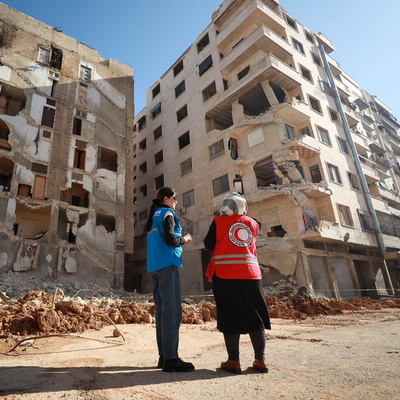 Une employée du HCR et une employée de la Croix-Rouge sont debout devant un bâtiment détruit.
