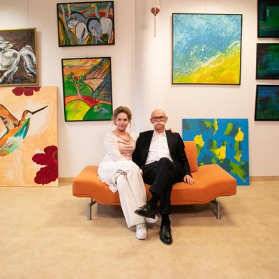 A woman and a man pose on an orange sofa, surrounded by art
