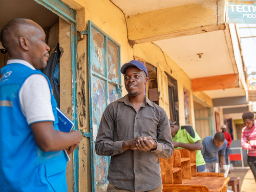 Two men, one in a UNHCR vest and one without, stand talking outside a row of buildings