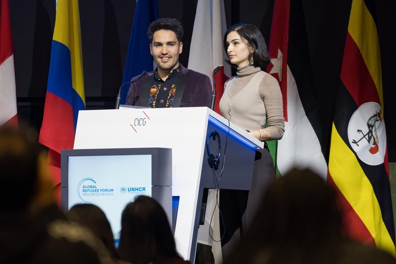 Two people speak from a podium with UNHCR and Global Refugee Forum branding, country flags lined up behind them.