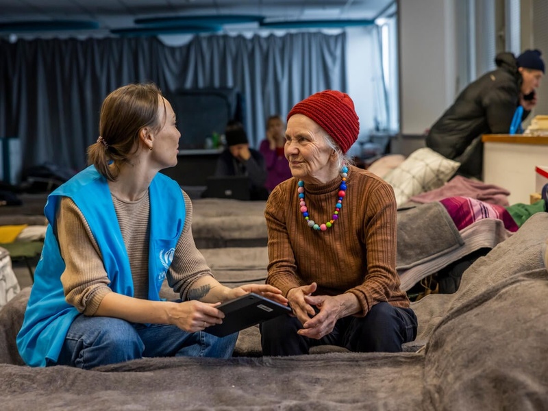 UNHCR staff member discussing with an older woman, both sitting on a bed.
