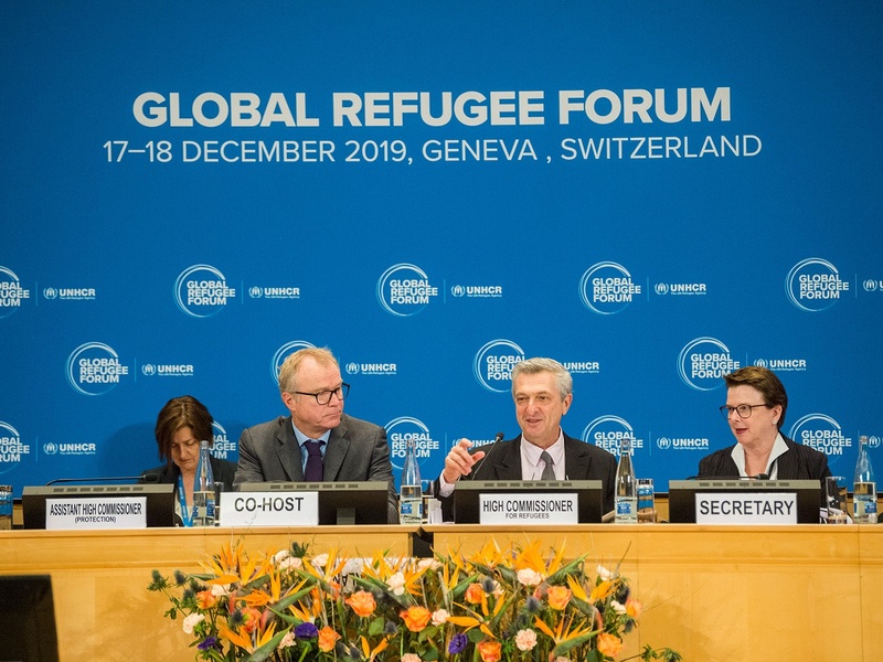 Three people (from right to left: Co-host, High Commissioner for Refugees and Secretary) sit at a podium at the Global Refugee Forum in December 2019, with one person (Assistant High Commissioner (Protection)) in the background.