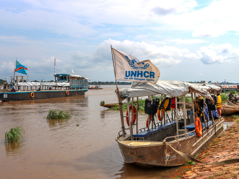 Wooden boats float on a wide, muddy river. One bears a UNHCR flag.