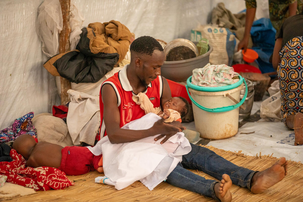 A man sitting on the floor inside a shelter holds his child on his lap while another baby sleeps next to him