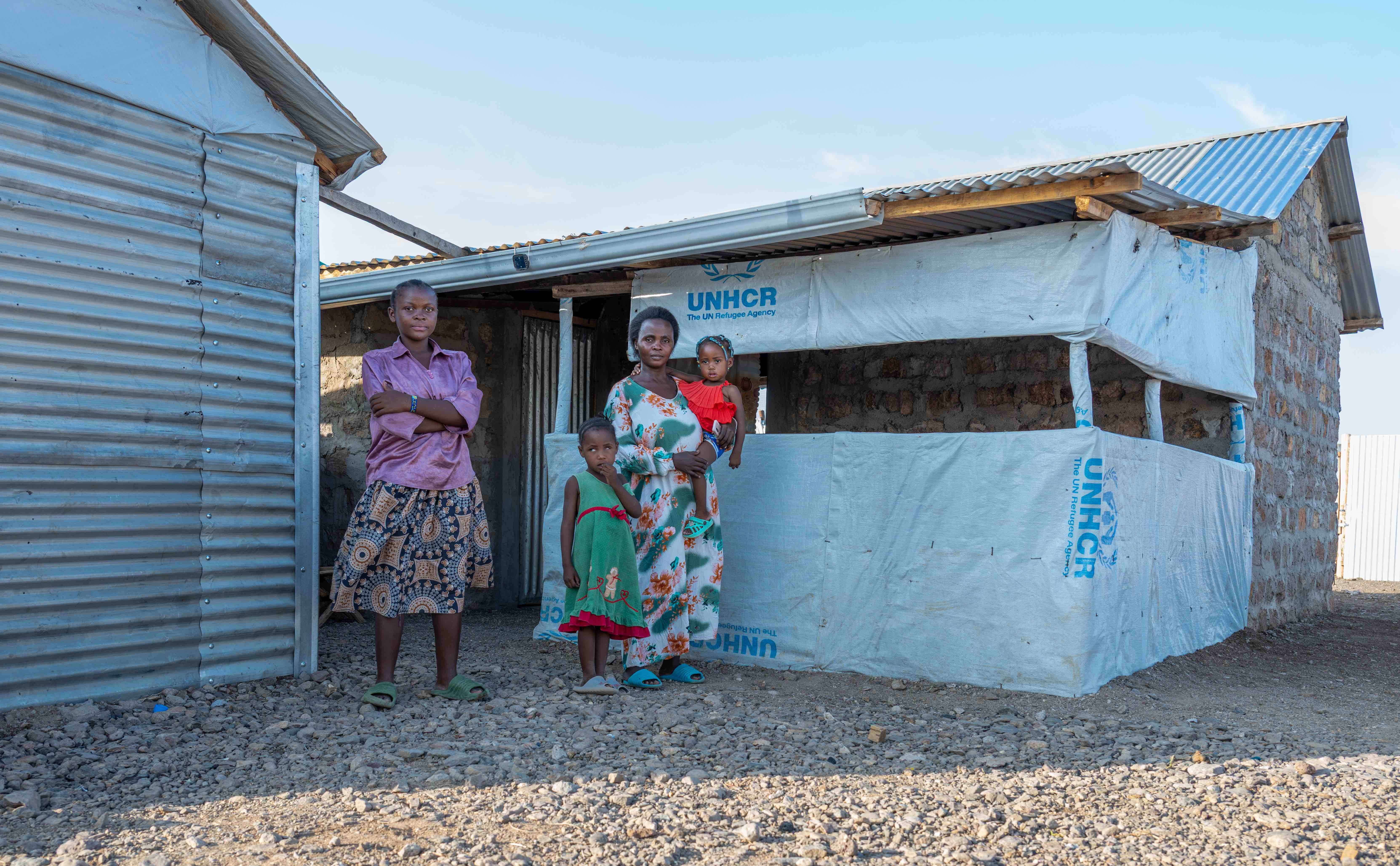 Sofia stands outside the shelter she built with support from UNHCR, alongside her daughters, Feza, 13, and Sakina, 5, while carrying her two-year-old daughter, Apolina.