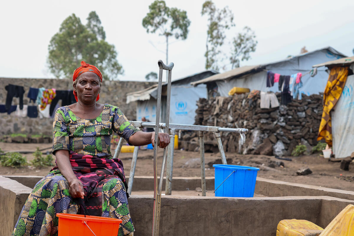 An elderly woman sits next to a communal tap holding a crutch in one hand and a bucket in the other.