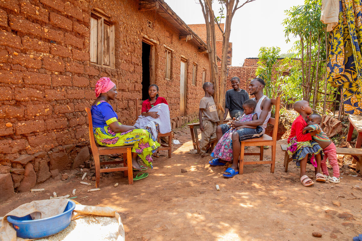 Two women, a man and children sit on wooden chairs in front of a mud house.