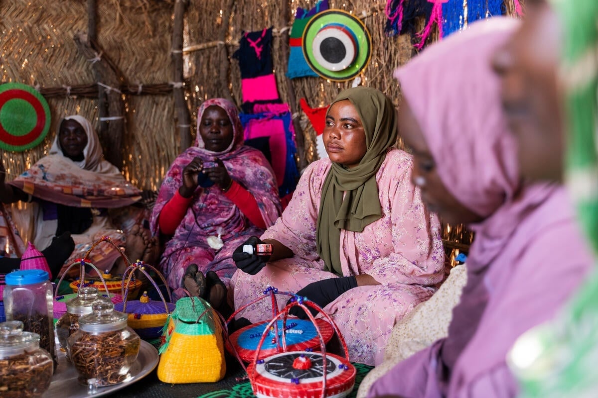 Women sit on the floor of a shelter showing off crafts they have made.