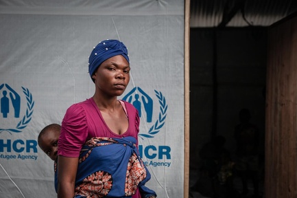 A woman carrying her baby on her back stands in front of a UNHCR shelter.