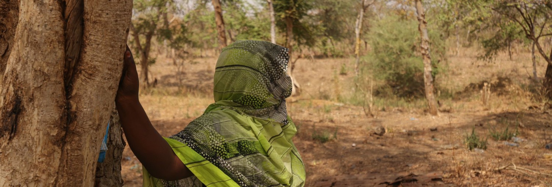 A woman leans against a tree looking out into the scrubland.