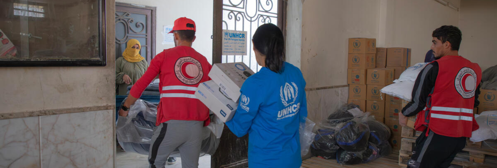 UNHCR and Syrian Arab Red Crescent staff carry boxes and bags of supplies out of a room.