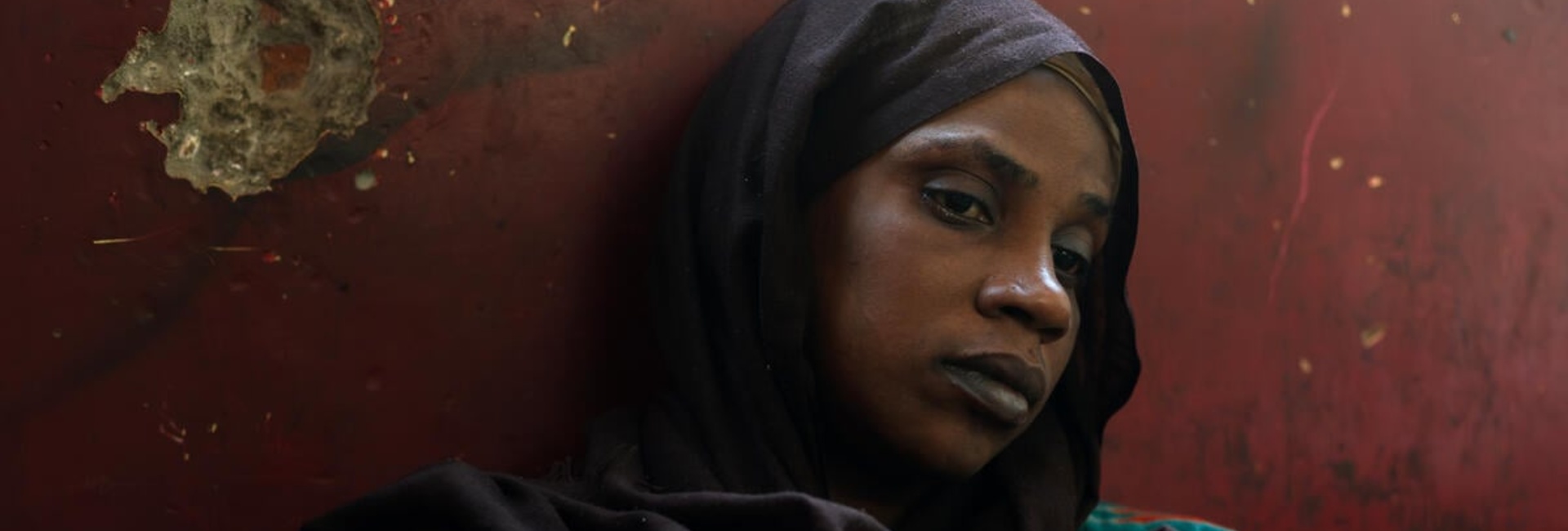 A woman in a colourful dress and a black headscarf sits on the floor leaning against a red-painted, pockmarked wall
