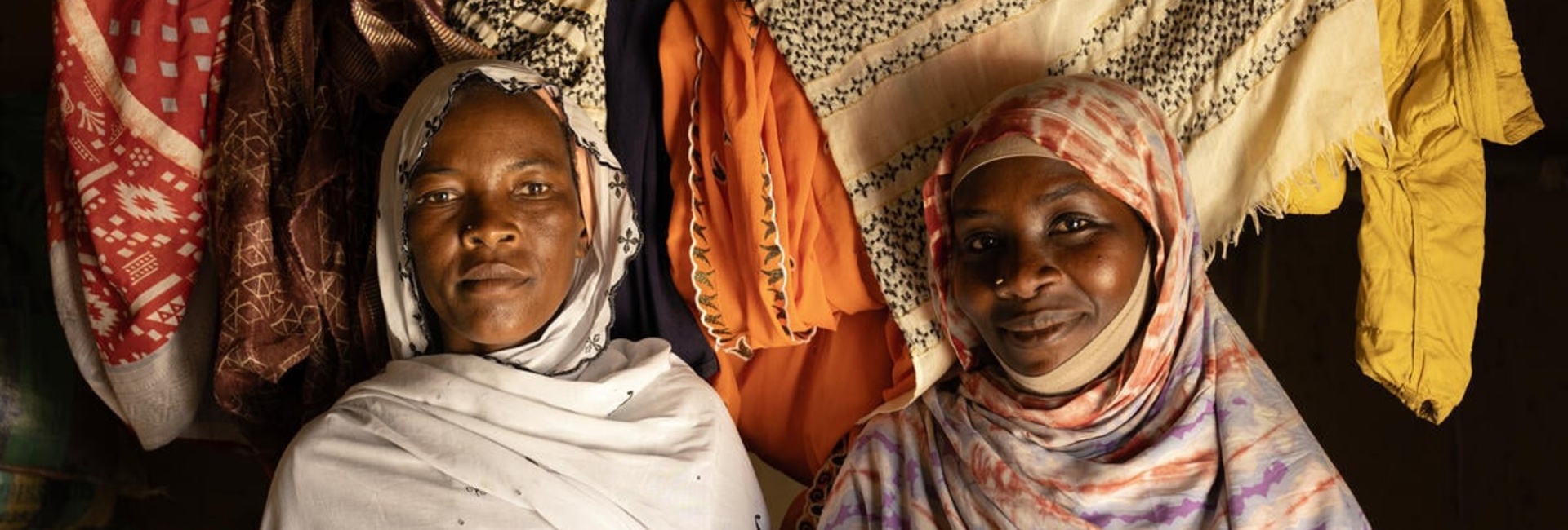 Two women stand next to each other inside a room with washing hanging on a line behind them