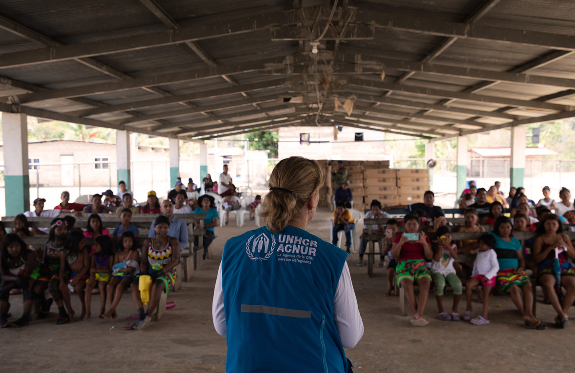 A woman in a branded UNHCR vest addresses a group of people in an open-sided building.