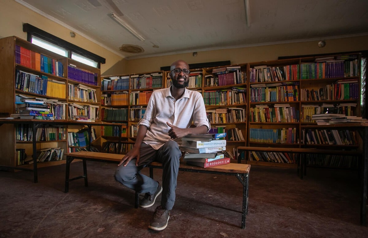 A man sits in a library, surrounded by books and leaning on a pile of books