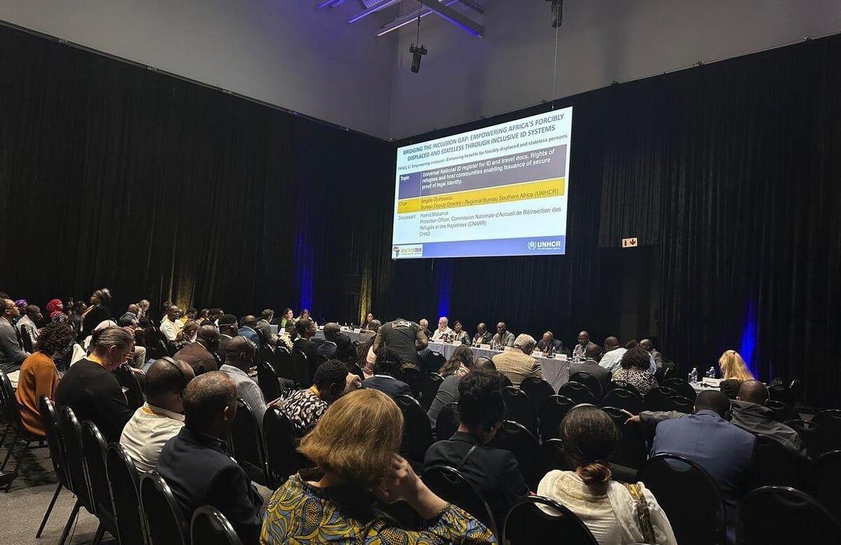 A large group of attendees at a conference look at a large screen with a PowerPoint presentation