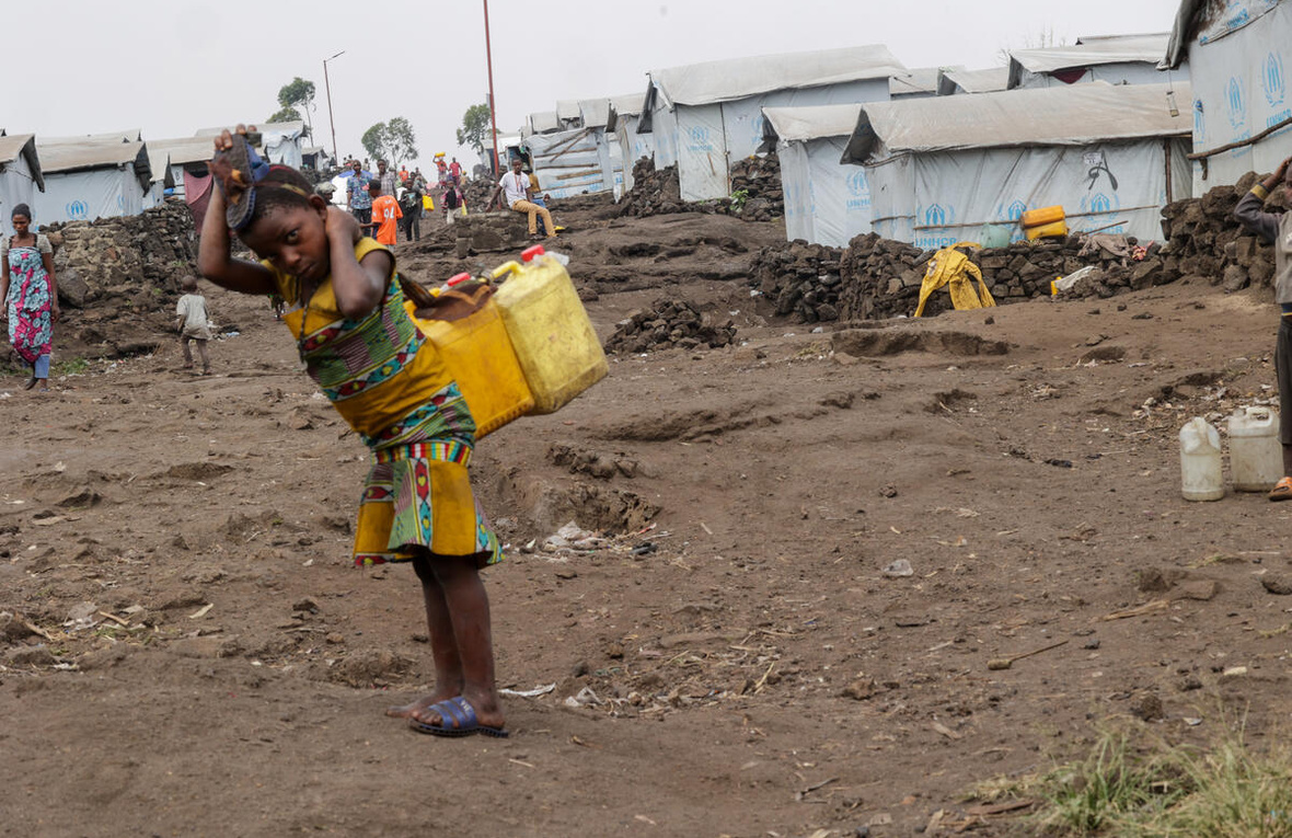 A young girl with two jerry cans on her back stands in front of a row of tents covered in tarpaulin.