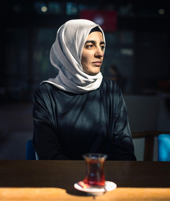 Aicha is surrounded by shadows with her face lit up by the sun as she looks off to the side, a cup of tea on a table in front of her