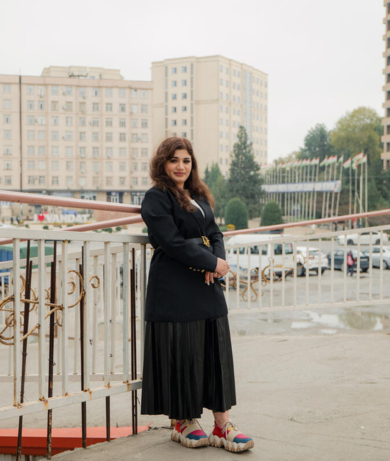 A young woman pictured leaning against a railing, with a carpark and tall buildings in the background
