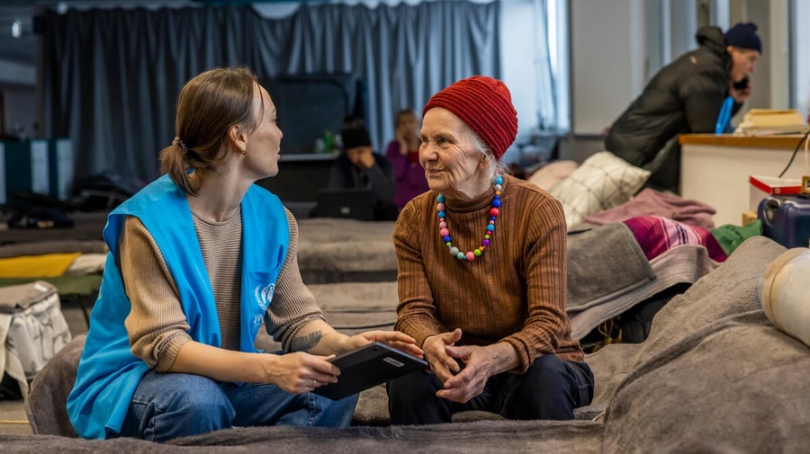 UNHCR staff member discussing with an older woman, both sitting on a bed.
