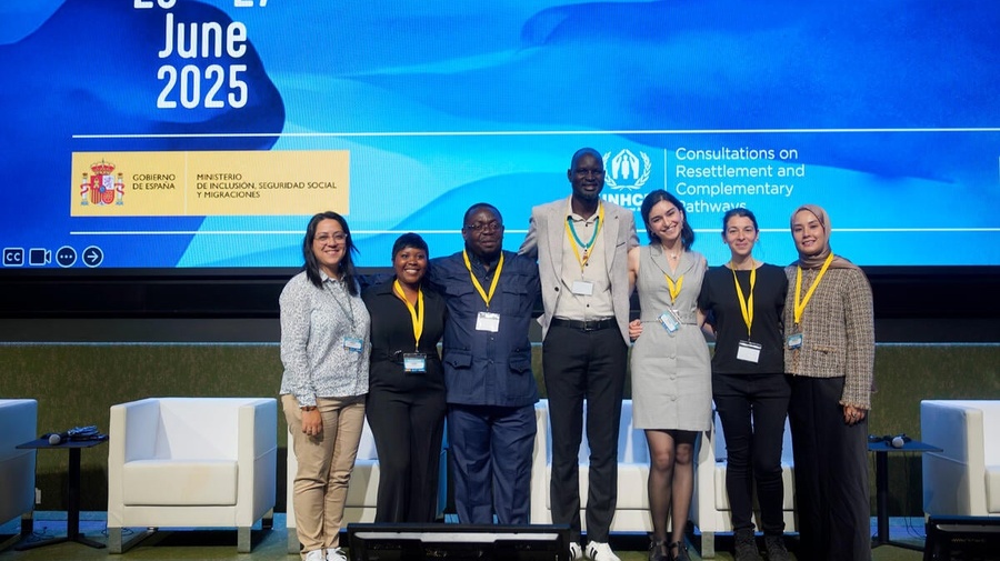 A group of 7 smiling people stand in front of a huge screen, displaying Consultations on Resettlement and Complementary Pathways, 25-27 June 2025 and a logo from the Spanish Government.