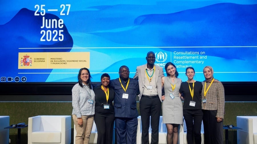 A group of 7 smiling people stand in front of a huge screen, displaying Consultations on Resettlement and Complementary Pathways, 25-27 June 2025 and a logo from the Spanish Government.