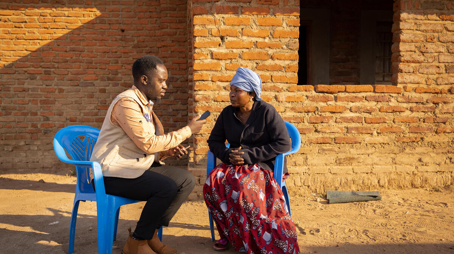A man interviews a woman outside on two plastic chairs, in front of a brick building