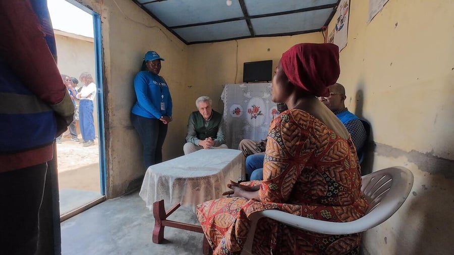 A woman seated on a plastic chair speaks to people seated and standing around a table inside a shelter
