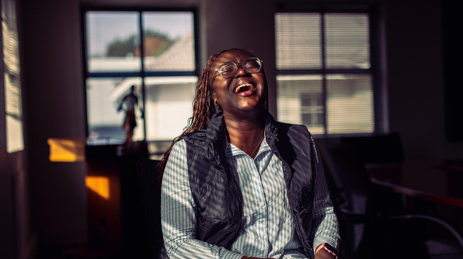 A woman sitting indoors in dappled light, with windows behind her, opens her mouth in laughter