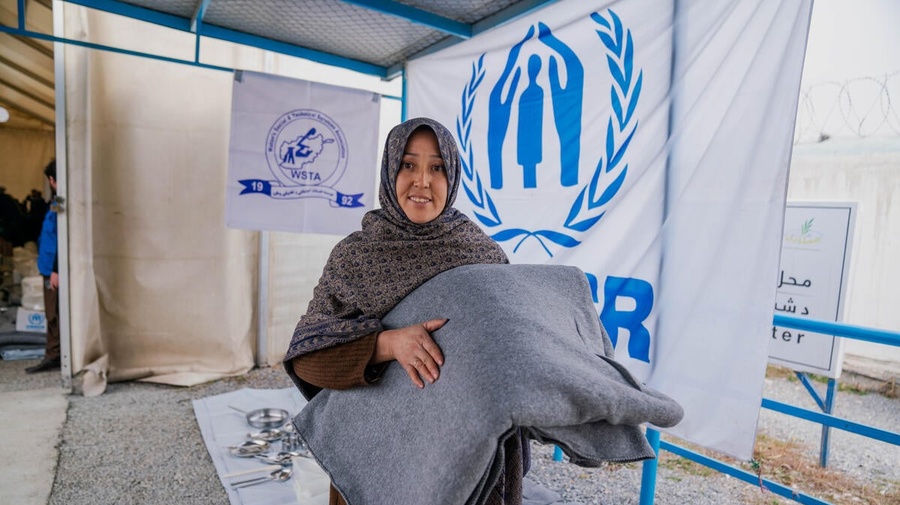 A woman smiles at the camera, holding some blankets. Behind her are other supplies like saucepans and blankets, and the logos of both UNHCR and WSTA.