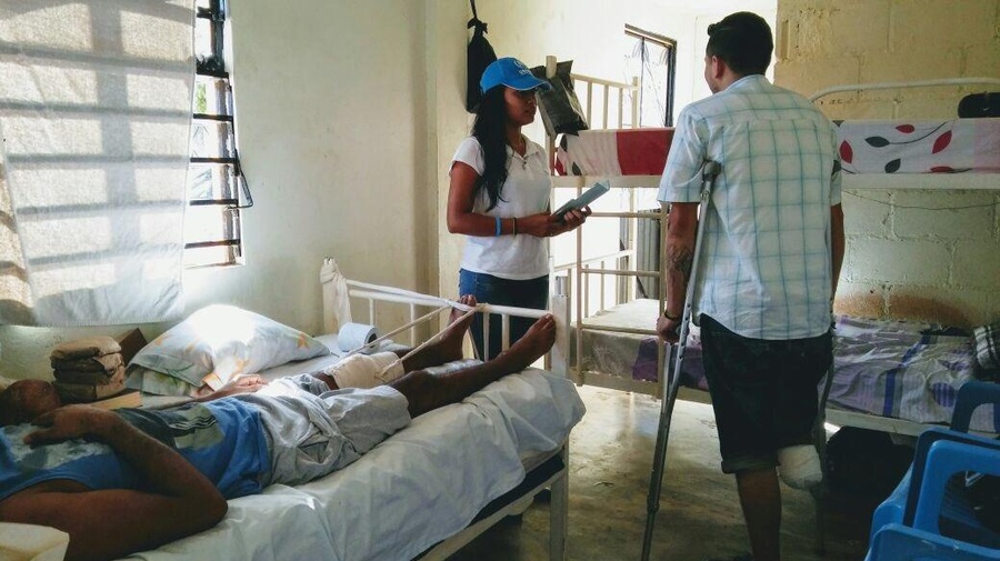 A man with an amputated leg stands with crutches and talks to an official. They are in a room with beds in it.