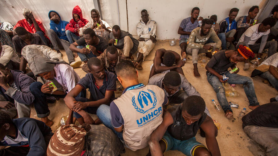 A group of disembarked refugees and migrants sit on the floor, joined by a UNHCR staff member wearing a tan UNHCR vest with blue logo.