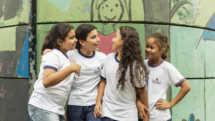 UNHCR UN Refugee Agency - How we do resettlement - ALT text: Four young girls enjoy chatting to each other while standing outside their classroom at the Duque de Caxias Municipal School in the Glicerio neighborhood of downtown Sao Paulo in Brazil. 