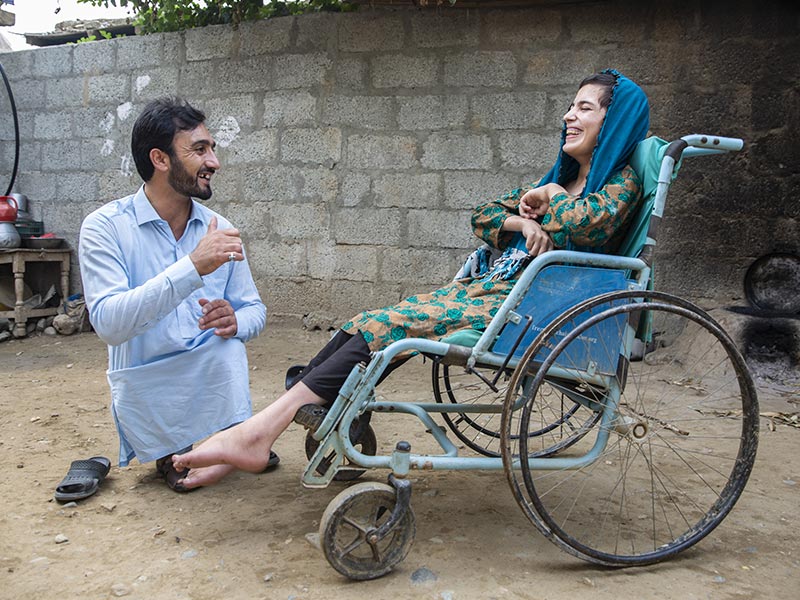 Two people talking and smiling, both wearing blue, one of them using a blue wheelchair.