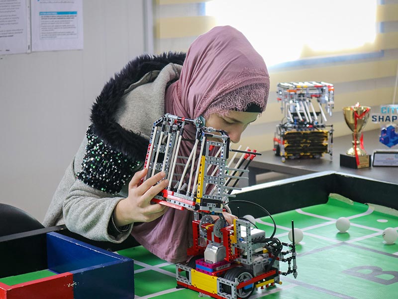 A girl wearing a pink headscarf and grey coat, bending over a table working on a robot. She looks very focused.