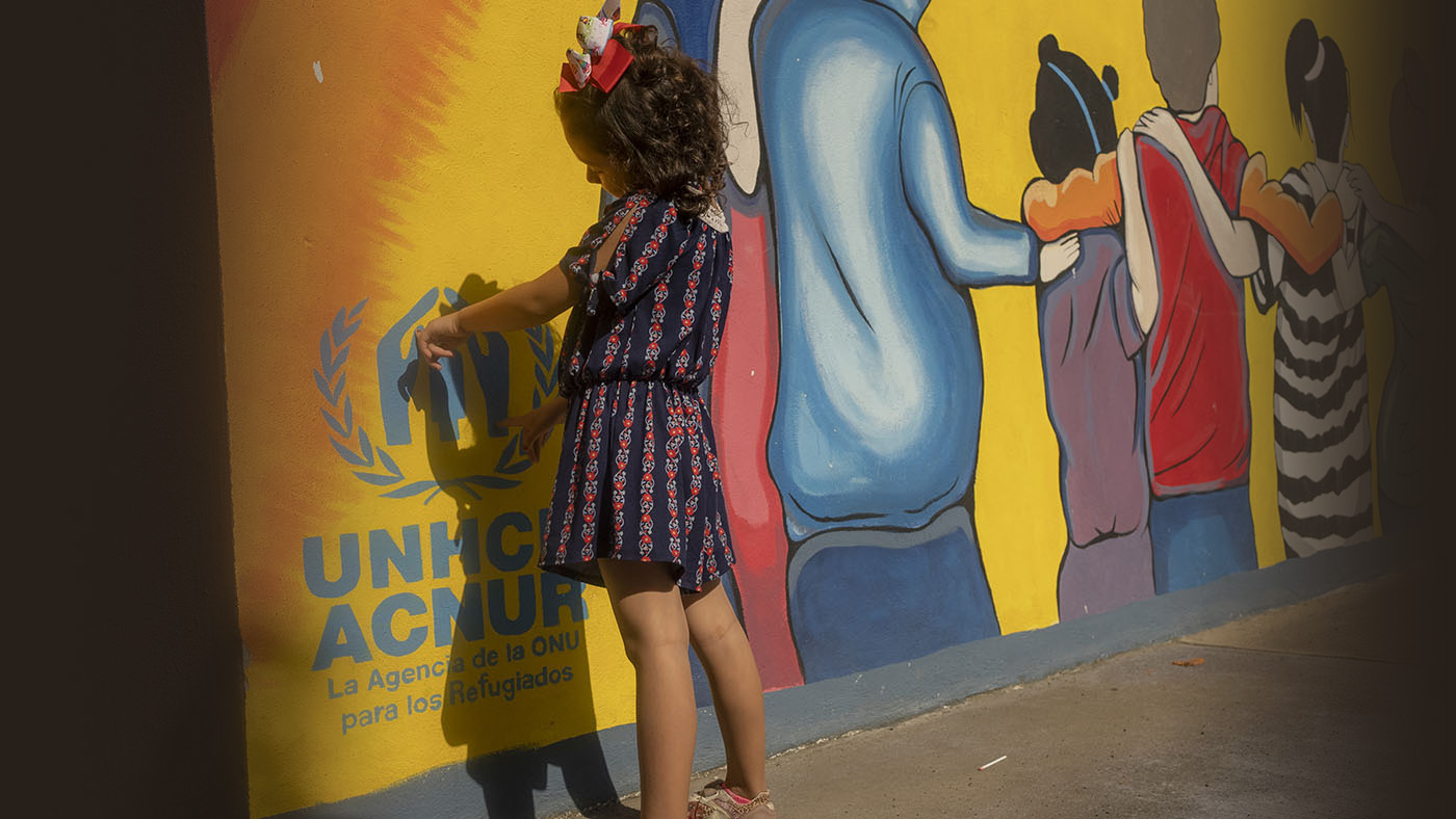 A young girl stands in front of a wall covered with murals while playing with her shadow